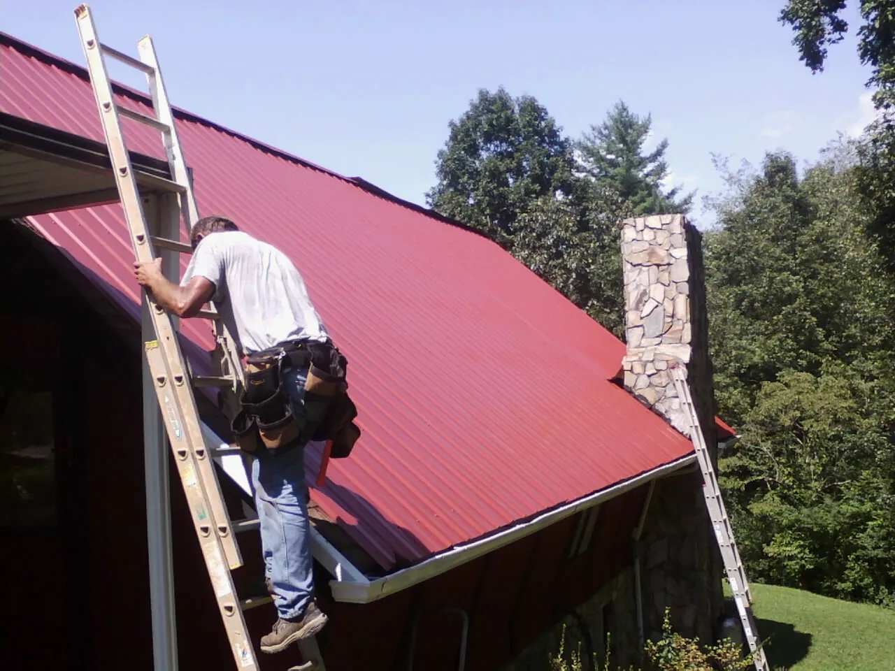 Roofer on a ladder beside a red metal roof and stone chimney — Soffit & Fascia Repair by Roofing Brothers in Rawlins